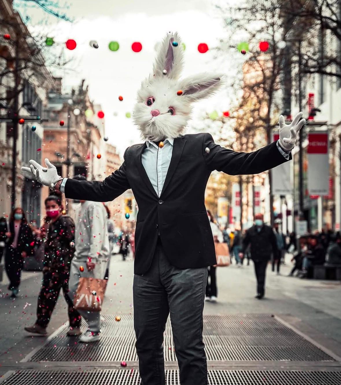 A person in a suit and a full rabbit head, arms wide on a shopping street as confetti falls.
